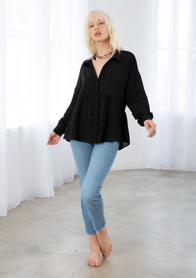 [Color: Black] A model wearing a sheer bohemian black button up shirt. With voluminous long sleeves, a button cuff, a tiered body, and a lace trim detail along the shoulder. 