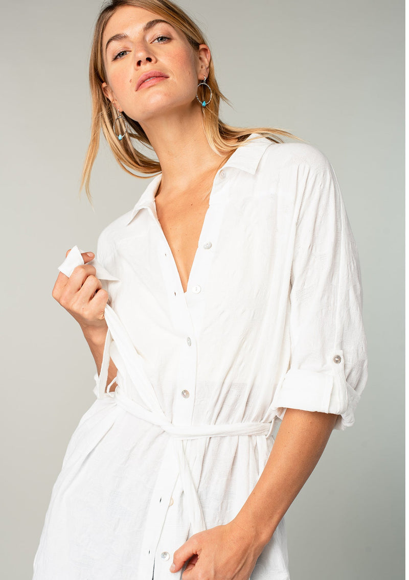 [Color: White] A close up front facing image of a blonde model wearing a bohemian white cotton tunic shirt in a textured jacquard. With long rolled sleeves and a button tab closure, a collared neckline, a button front, a self tie waist belt, side vents, a round hemline, and a back yoke detail. 