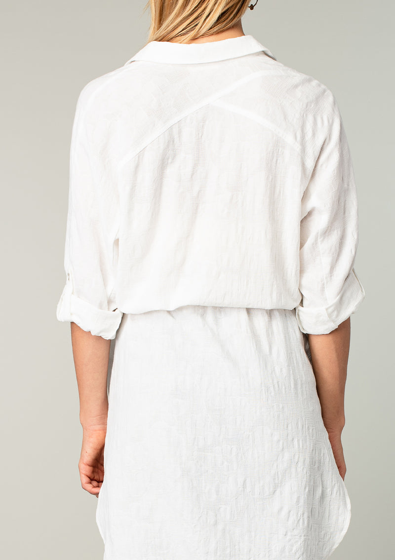 [Color: White] A back facing image of a blonde model wearing a bohemian white cotton tunic shirt in a textured jacquard. With long rolled sleeves and a button tab closure, a collared neckline, a button front, a self tie waist belt, side vents, a round hemline, and a back yoke detail. 