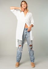 [Color: White] A front facing image of a blonde model wearing a bohemian white cotton tunic shirt in a textured jacquard. With long rolled sleeves and a button tab closure, a collared neckline, a button front, a self tie waist belt, side vents, a round hemline, and a back yoke detail. 