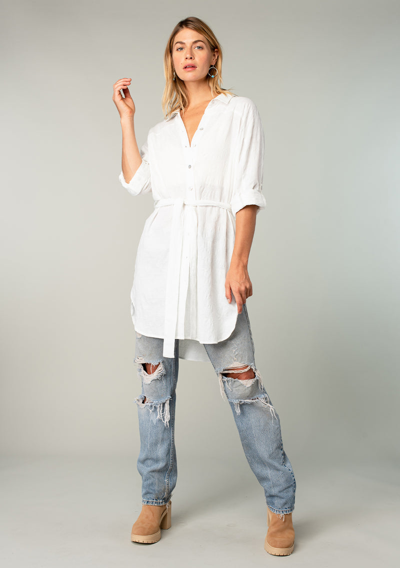 [Color: White] A full body front facing image of a blonde model wearing a bohemian white cotton tunic shirt in a textured jacquard. With long rolled sleeves and a button tab closure, a collared neckline, a button front, a self tie waist belt, side vents, a round hemline, and a back yoke detail. 