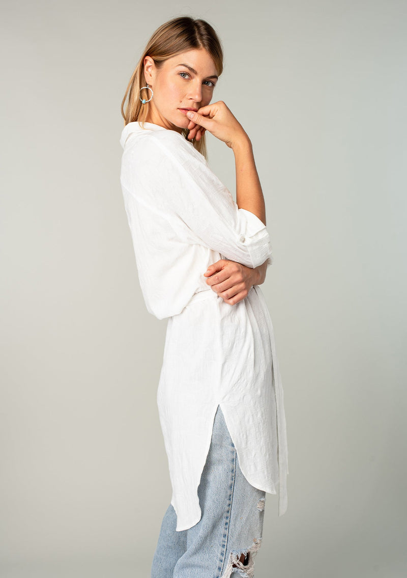 [Color: White] A side facing image of a blonde model wearing a bohemian white cotton tunic shirt in a textured jacquard. With long rolled sleeves and a button tab closure, a collared neckline, a button front, a self tie waist belt, side vents, a round hemline, and a back yoke detail. 
