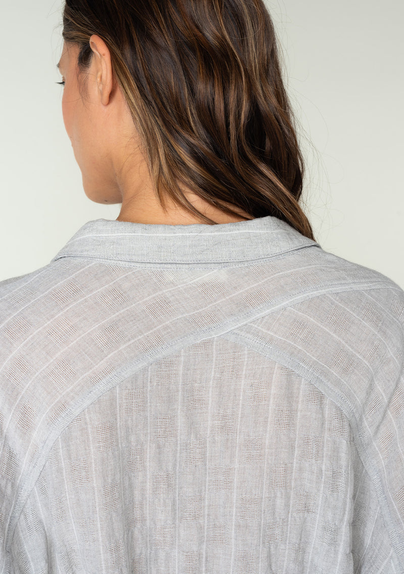 [Color: Heather Grey] A back facing image of a brunette model wearing a sheer cotton heather grey tunic top in a jacquard stripe. With long rolled sleeves, a button front, a collared neckline, an extra long length, and a self tie waist belt. 