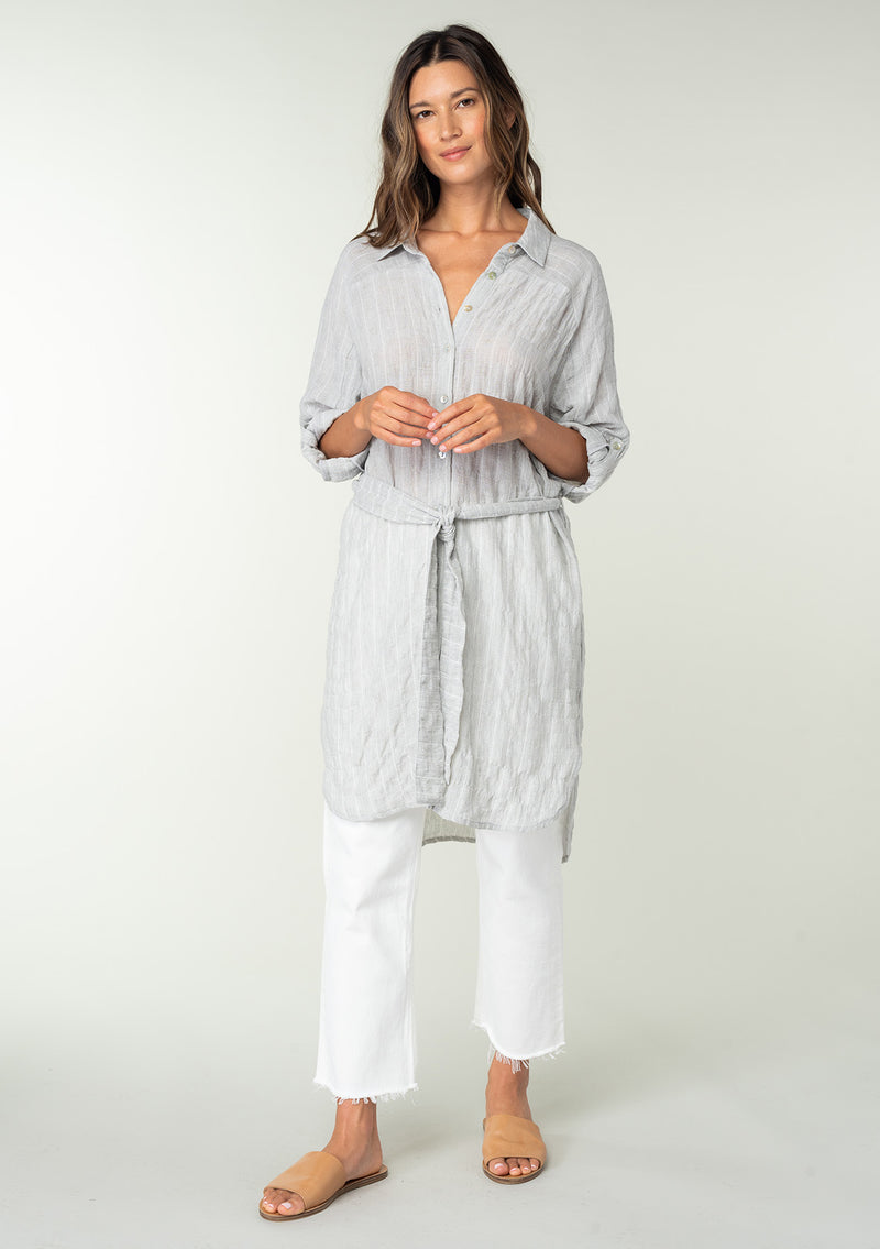 [Color: Heather Grey] A full body front facing image of a brunette model wearing a sheer cotton heather grey tunic top in a jacquard stripe. With long rolled sleeves, a button front, a collared neckline, an extra long length, and a self tie waist belt. 