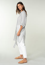 [Color: Heather Grey] A side facing image of a brunette model wearing a sheer cotton heather grey tunic top in a jacquard stripe. With long rolled sleeves, a button front, a collared neckline, an extra long length, and a self tie waist belt. 