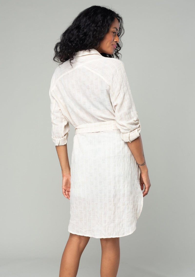 [Color: Oatmeal] A back facing image of a brunette model wearing a sheer cotton off white tunic top in a jacquard stripe. With long rolled sleeves, a button front, a collared neckline, an extra long length, and a self tie waist belt.