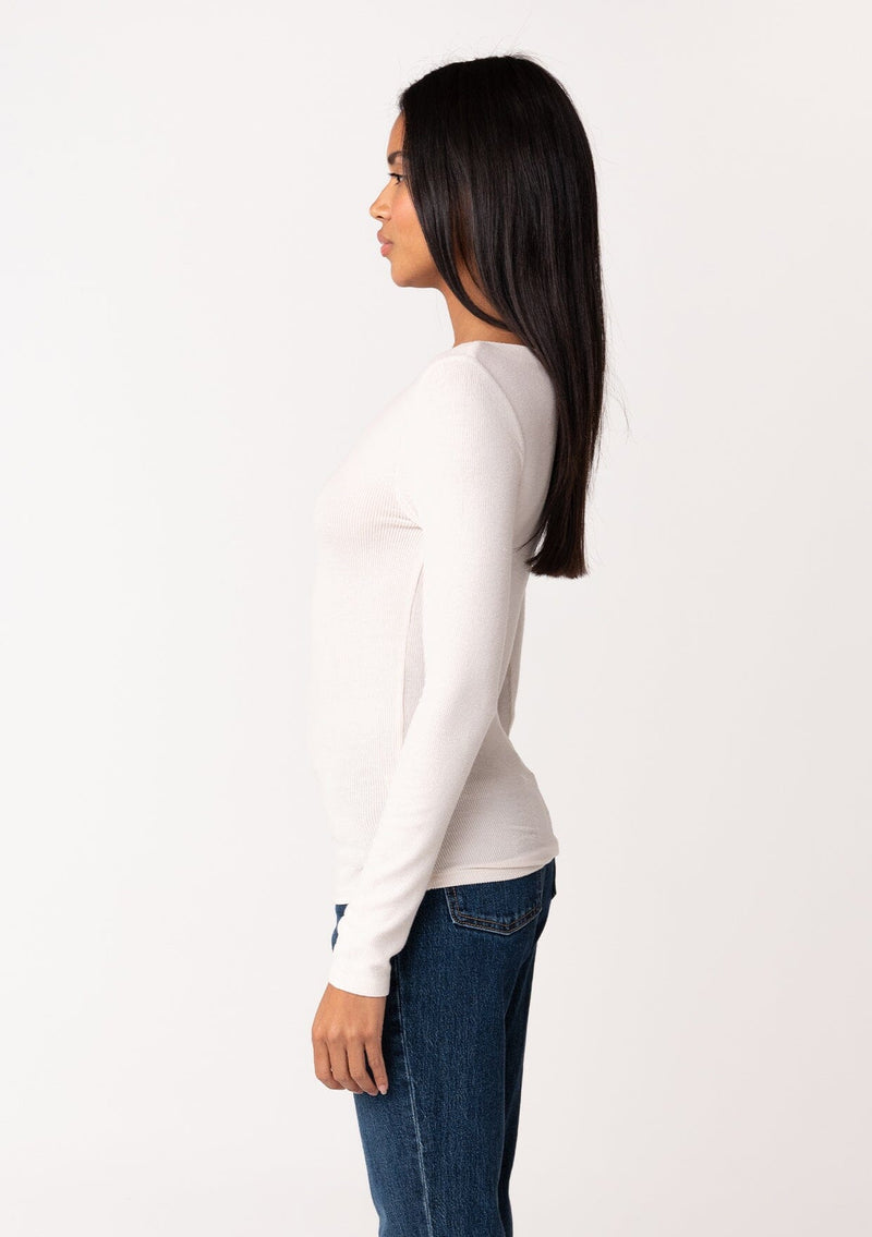 [Color: Nude] A side facing image of a brunette model wearing an off white bamboo micro ribbed long sleeve tee. Featuring a mock neckline and a stretchy slim fit.