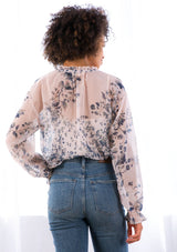 [Color: Light Peach] A model wearing a sheer peasant top in a vintage floral print. With a ruffled split neckline and long voluminous sleeves with a flounced wrist cuff.