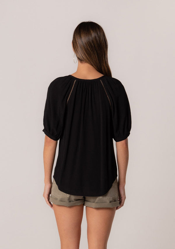 [Color: Black] A back facing image of a brunette model wearing a best selling button front blouse in black. With short puff sleeves, a button front, and a split v neckline with ties. 