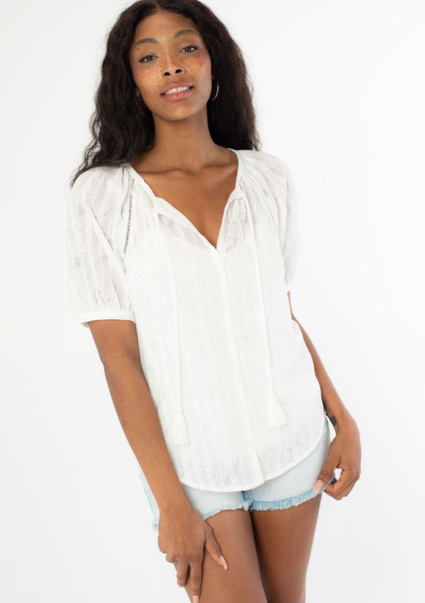 [Color: White] A front facing image of a black model wearing a white bohemian blouse with short puff sleeves, a self covered button front, and tassel ties. 