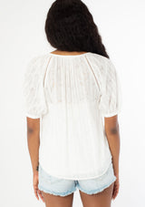 [Color: White] A back facing image of a black model wearing a white bohemian blouse with short puff sleeves, a self covered button front, and tassel ties. 