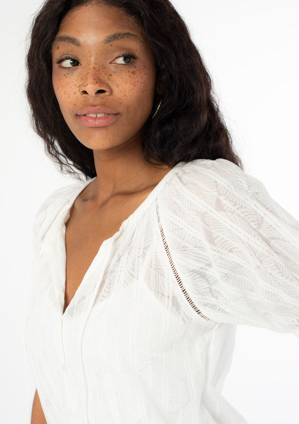 [Color: White] A close up side facing image of a black model wearing a white bohemian blouse with short puff sleeves, a self covered button front, and tassel ties. 