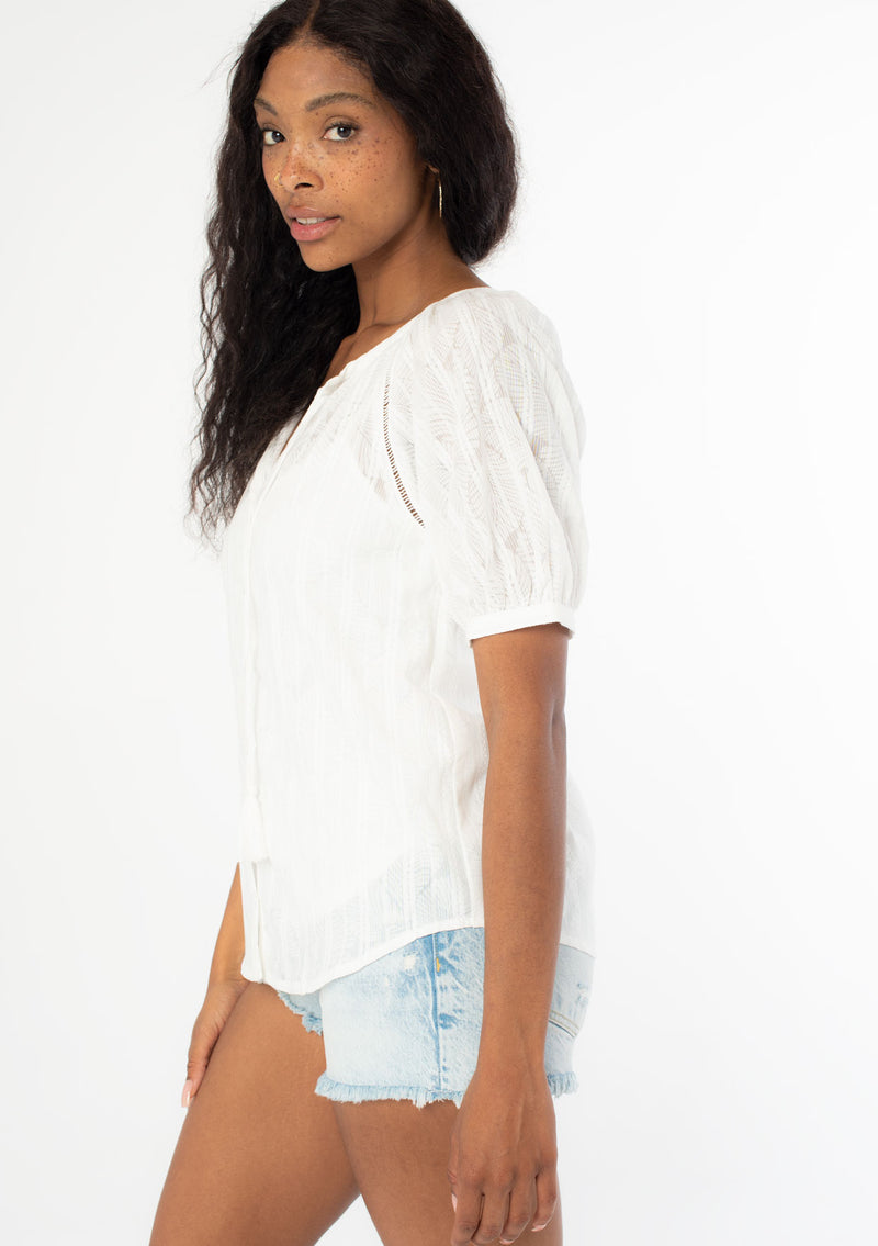 [Color: White] A side facing image of a black model wearing a white bohemian blouse with short puff sleeves, a self covered button front, and tassel ties. 