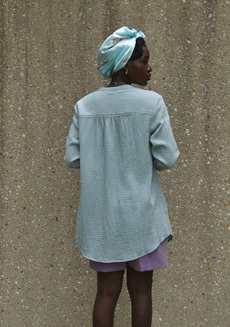 [Color: Dusty Teal] A woman wearing a textured airy gauze button front shirt. With a long tunic length, long rolled sleeves with a button tab, a single front pocket, and a banded collar with a v neckline.