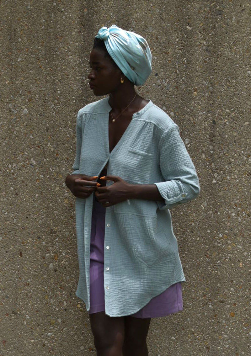 [Color: Dusty Teal] A woman wearing a textured airy gauze button front shirt. With a long tunic length, long rolled sleeves with a button tab, a single front pocket, and a banded collar with a v neckline.