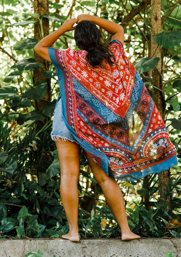 [Color: Blue/Brick] A model wearing a red and blue bohemian mid length kimono. Featuring half length kimono sleeves, side slits, an open front, and a multi colored pattern throughout.