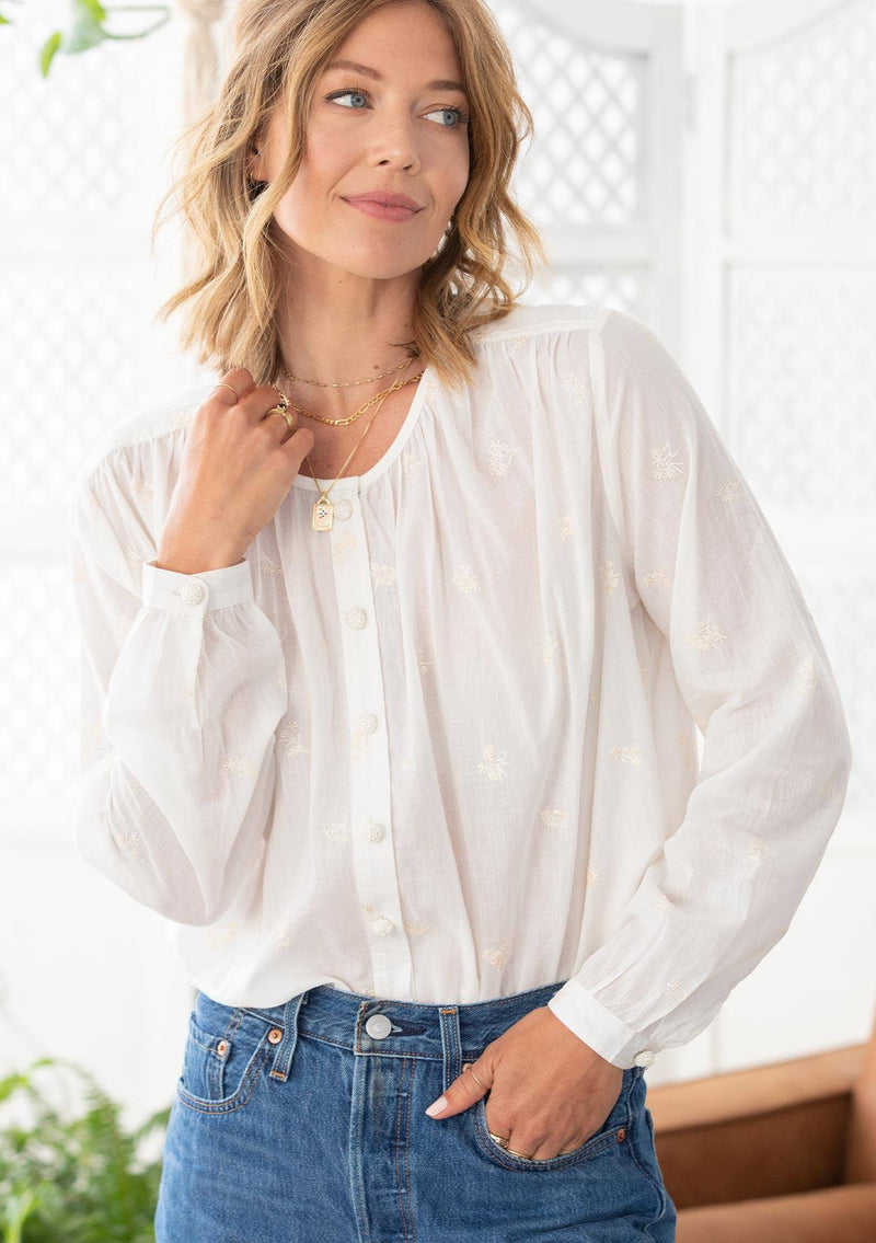 [Color: Cloud] A woman wearing a sheer cotton voile button front top. Featuring pretty floral embroidered details throughout, voluminous long sleeves with a button cuff closure, and a large button up front.