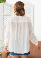 [Color: Cloud] A woman wearing a sheer cotton voile button front top. Featuring pretty floral embroidered details throughout, voluminous long sleeves with a button cuff closure, and a large button up front.