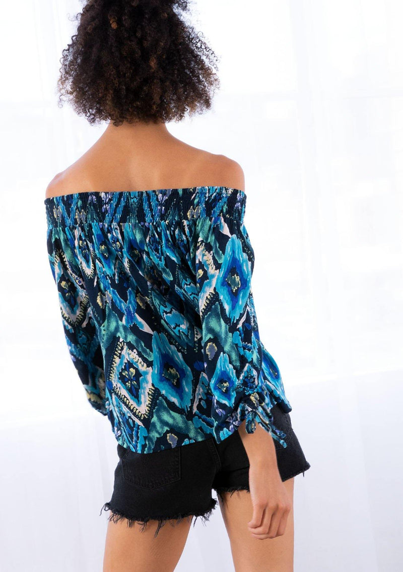 [Color: Navy/Teal] A model wearing a off the shoulder top in a teal watercolor geometric print. With a voluminous three quarter length sleeve with gathered tie detail, a smocked elastic neckline, and a billowy fit.