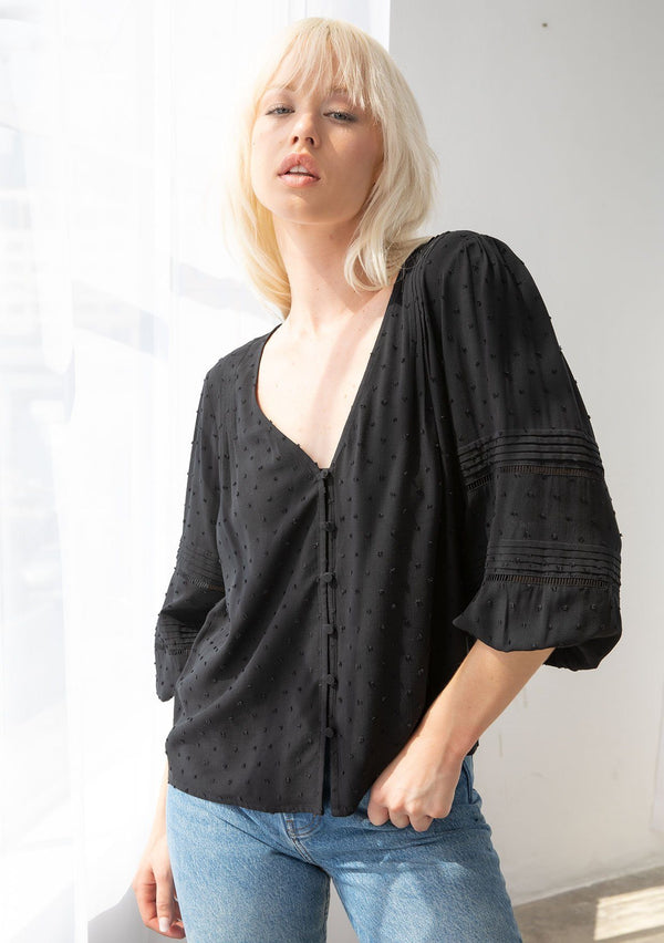 [Color: Black] A model wearing a classic bohemian black peasant top in a clip dot fabric. With three quarter voluminous sleeves, a self covered button front, a v neckline, and delicate pin tuck details throughout. 