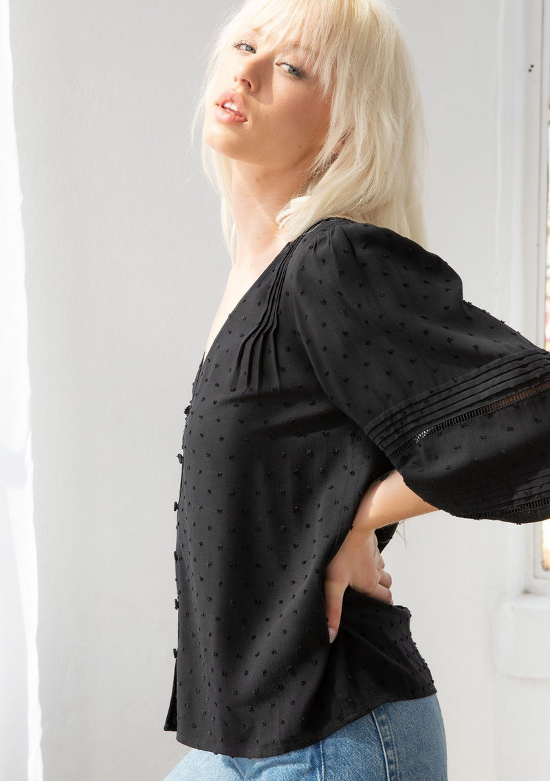 [Color: Black] A model wearing a classic bohemian black peasant top in a clip dot fabric. With three quarter voluminous sleeves, a self covered button front, a v neckline, and delicate pin tuck details throughout. 