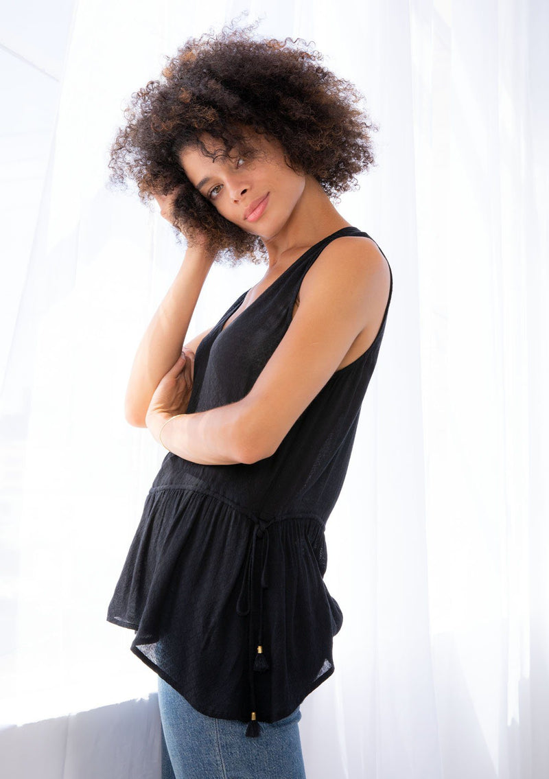 [Color: Black] A model wearing a black tank top in textured jacquard. With a v neckline, a drawstring waist, tassel tie side detail, and thicker tank top straps. 