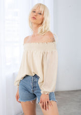[Color: Vanilla] A model wearing a bohemian vanilla cream off shoulder top in a sheer shadow stripe. With long voluminous sleeves, a smocked elastic wrist cuff, and a smocked elastic off shoulder neckline. 