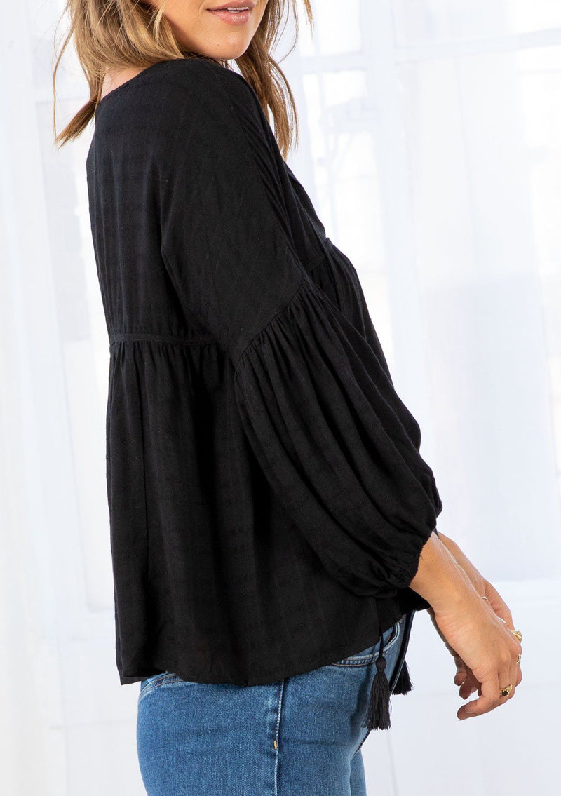 [Color: Black] A model wearing a slightly cropped classic black peasant top. Featuring a split v neckline with tassel ties, long voluminous balloon sleeves, and gathered details along the yoke. 