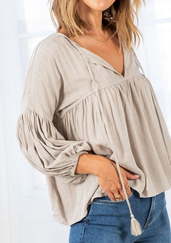 [Color: Stone] A model wearing a slightly cropped classic stone grey peasant top. Featuring a split v neckline with tassel ties, long voluminous balloon sleeves, and gathered details along the yoke. 
