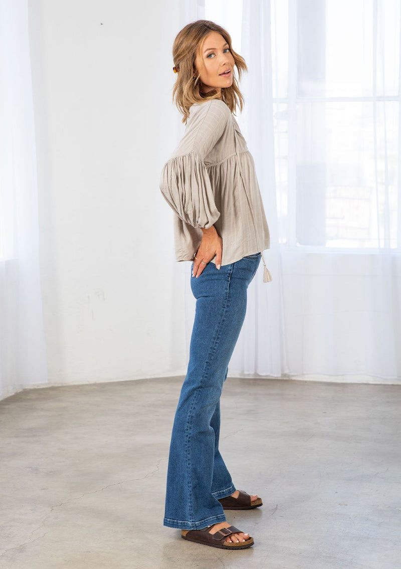 [Color: Stone] A model wearing a slightly cropped classic stone grey peasant top. Featuring a split v neckline with tassel ties, long voluminous balloon sleeves, and gathered details along the yoke. 