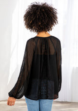 [Color: Black] A model wearing a sheer textured black peasant top with long voluminous sleeves, elastic wrist cuffs, a split v neckline with tassel ties, and delicate lace trim along the outer sleeve and center back. 