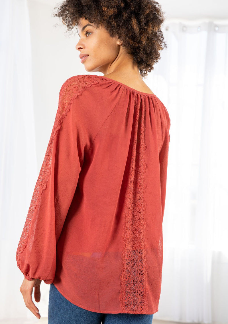 [Color: Cayenne] A model wearing a sheer textured cayenne red peasant top with long voluminous sleeves, elastic wrist cuffs, a split v neckline with tassel ties, and delicate lace trim along the outer sleeve and center back. 