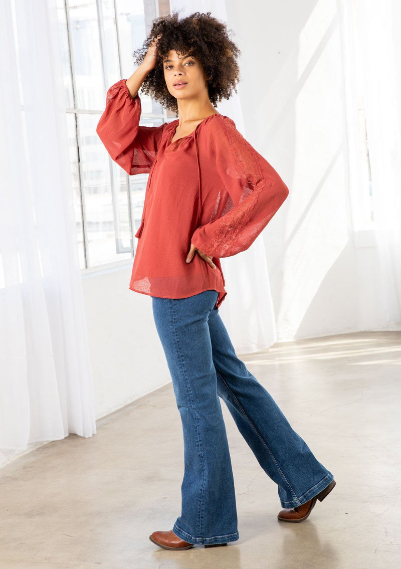 [Color: Cayenne] A model wearing a sheer textured cayenne red peasant top with long voluminous sleeves, elastic wrist cuffs, a split v neckline with tassel ties, and delicate lace trim along the outer sleeve and center back. 