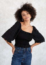 [Color: Black] A model wearing a cropped black bohemian top with a smocked bodice, a ruffle trimmed square neckline, a ruffled hemline, and a scalloped edge trimmed half length sleeve.
