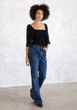 [Color: Black] A model wearing a cropped black bohemian top with a smocked bodice, a ruffle trimmed square neckline, a ruffled hemline, and a scalloped edge trimmed half length sleeve.
