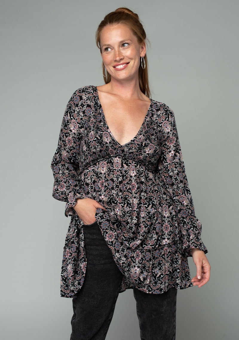 [Color: Black/Rose] A front facing image of a red headed model wearing a black and pink paisley print tunic top. A vintage inspired top with long sleeves, a smocked bodice, an empire waist, and a deep v neckline. 