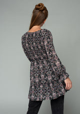 [Color: Black/Rose] A back facing image of a red headed model wearing a black and pink paisley print tunic top. A vintage inspired top with long sleeves, a smocked bodice, an empire waist, and a deep v neckline. 