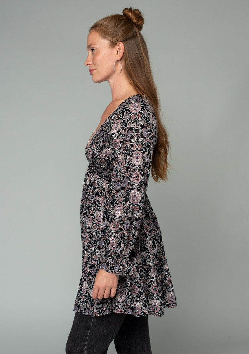 [Color: Black/Rose] A side facing image of a red headed model wearing a black and pink paisley print tunic top. A vintage inspired top with long sleeves, a smocked bodice, an empire waist, and a deep v neckline. 