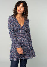 [Color: Navy/Tan] A half body front facing image of a brunette model wearing a navy blue and tan paisley print tunic top. A vintage inspired top with long sleeves, a smocked bodice, an empire waist, and a deep v neckline.
