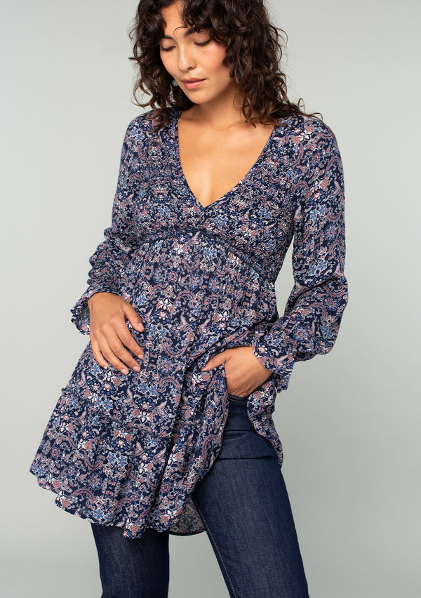 [Color: Navy/Tan] A front facing image of a brunette model wearing a navy blue and tan paisley print tunic top. A vintage inspired top with long sleeves, a smocked bodice, an empire waist, and a deep v neckline.