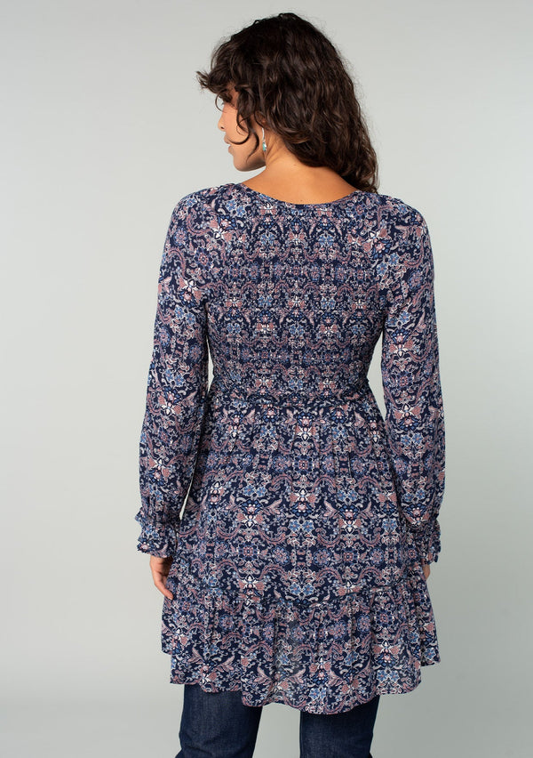 [Color: Navy/Tan] A back facing image of a brunette model wearing a navy blue and tan paisley print tunic top. A vintage inspired top with long sleeves, a smocked bodice, an empire waist, and a deep v neckline.