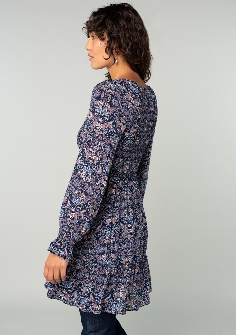 [Color: Navy/Tan] A side facing image of a brunette model wearing a navy blue and tan paisley print tunic top. A vintage inspired top with long sleeves, a smocked bodice, an empire waist, and a deep v neckline.
