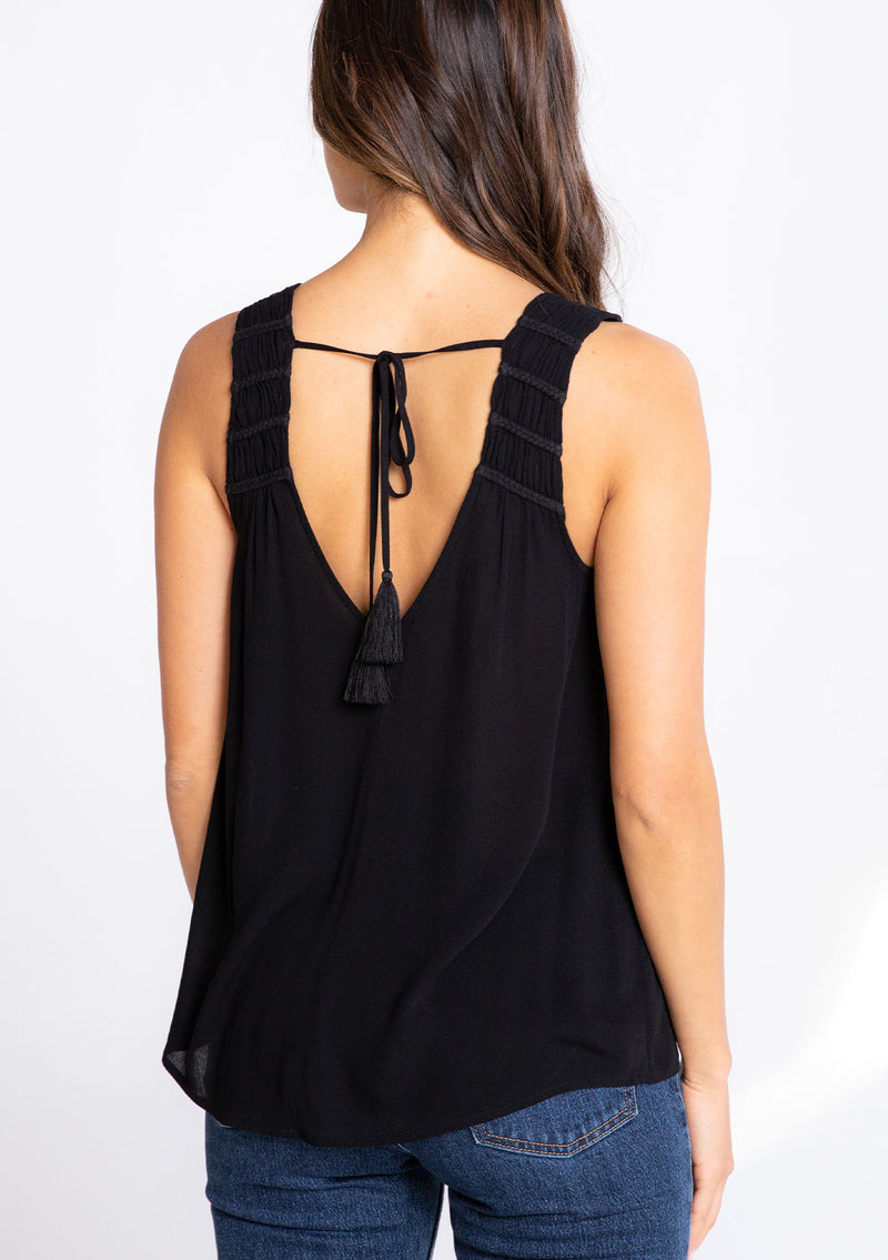 [Color: Black] A model wearing a flowy black bohemian tank top with a v neckline in front and back and braided trim details. 