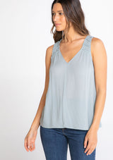 [Color: Blue Fog] A model wearing a flowy light blue bohemian tank top with a v neckline in front and back and braided trim details. 
