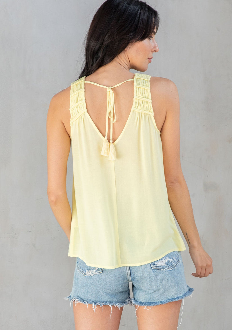 [Color: Yellow] A model wearing a flowy yellow bohemian tank top with a v neckline in front and back and braided trim details. 