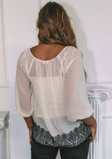 [Color: Gardenia/Black] A model wearing an ethereal sheer white chiffon blouse with black eyelash lace trim hemline.