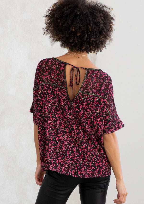[Color: Black/Raspberry] A model wearing a black and red ditsy floral chiffon top. With short cuffed sleeves, a back keyhole detail with tie closure, gold thread trim throughout, and a boxy, relaxed fit. 