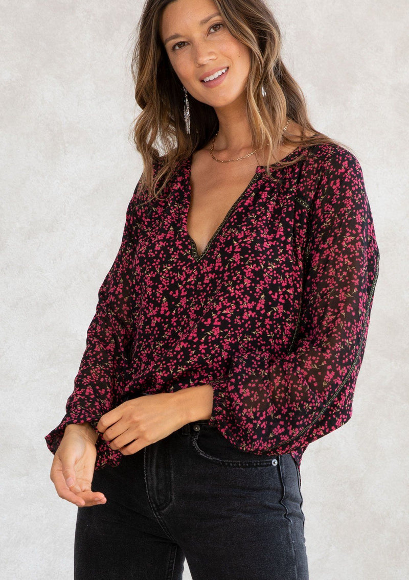 [Color: Black/Raspberry] A model wearing a holiday inspired black and red ditsy floral print chiffon peasant top. With gold thread trim throughout, long voluminous raglan sleeves, and a split v neckline. 