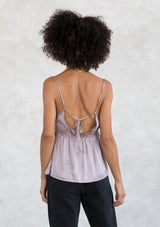[Color: Lilac Mist] A model wearing an elevated lilac purple silky camisole with strappy back detail. With a v neckline, adjustable spaghetti straps, and a flattering relaxed silhouette. 