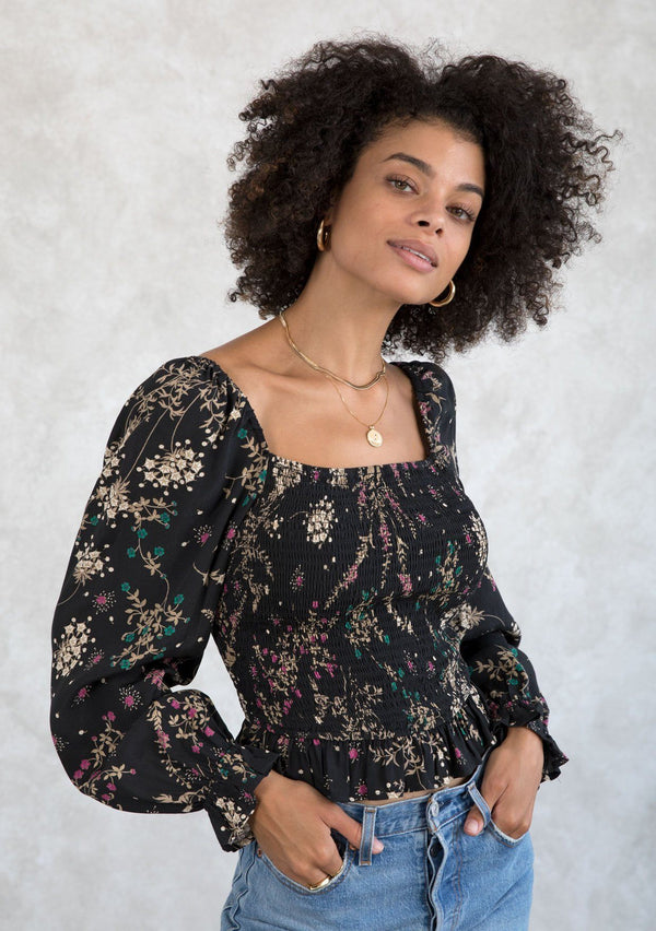 [Color: Black/Jade] A model wearing a black and green floral print bohemian top. With a smocked bodice, long voluminous sleeves, and a ruffle trimmed cropped length. 